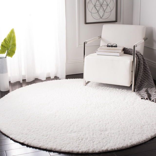 SAFAVIEH August Carlene Solid Plush Shag Area Rug in white, round accent rug with plush texture on dark hardwood floor. Positioned in a bright room with sheer curtains, under a modern white armchair with chrome arms, gray throw, and stacked books with glasses. Suitable for adding softness and style to living spaces.