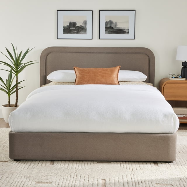 Better Homes & Gardens Juliet Queen Platform Bed in sand with a low-profile upholstered wood frame and rounded tufted headboard, shown front-facing with white bedding and a tan leather accent pillow, placed in a modern bedroom with a potted plant and wooden nightstand featuring a lamp and glasses, suitable for memory foam or hybrid mattresses.