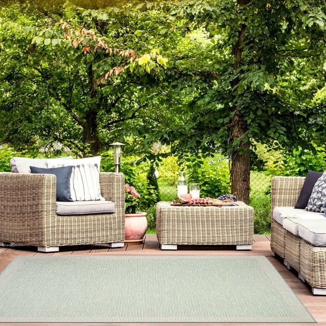 ECARPET Outdoor Area Rug for Patio, Deck, Backyard, Camping, Water Proof Carpet, Veranda Classic Collection in light green woven pattern on a wooden deck with wicker cushioned armchair and sofa. The backyard setting includes trees and plants with a wicker ottoman holding a tray with a bottle and glasses, ideal for outdoor living spaces and gatherings.