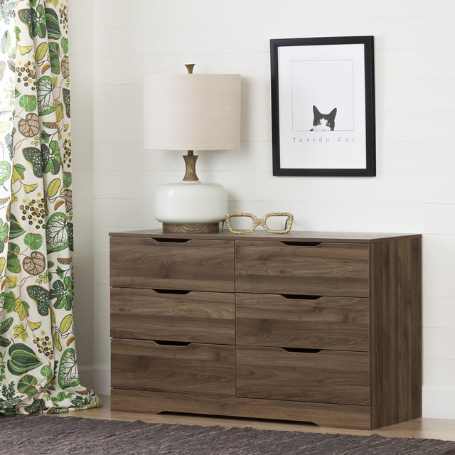 South Shore Holland 6-Drawer Dresser for Adult in natural walnut finish with six spacious drawers featuring cutout handles and a curved kick plate. The dresser is styled with a white ceramic lamp with beige shade and decorative glasses on top, set against a white shiplap wall with a framed 'Tuxedo Cat' print. A green and white botanical curtain and textured rug complete the bedroom setting.