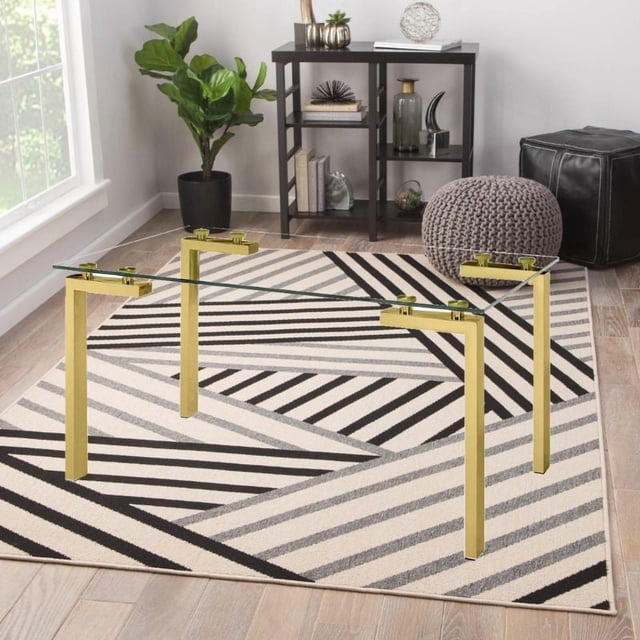 TAKE ME HOME RECTANGLE TABLE G50" with clear tempered glass top and gold stainless steel legs, placed on a geometric black, white, and gray rug in a bright room with light wood flooring, near a black shelving unit, potted plant, gray knitted pouf, and black leather ottoman. Suitable for dining or desk use.