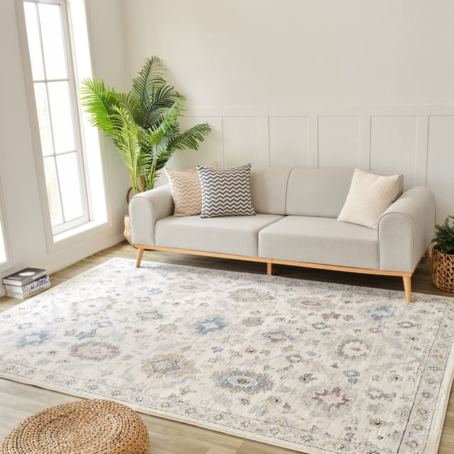 Rug Branch Nomad Classic Indoor Area Rug in beige cream with boho floral and geometric patterns in muted blue, green, and purple tones, placed on a light wood floor in a bright living room featuring a modern light grey sofa with chevron and textured pillows and woven plant baskets, ideal for living room, bedroom, dining room, or kitchen.
