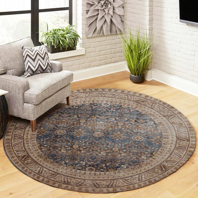 ECARPET Gianna Traditional Area Rug, round with a deep blue patterned center and neutral beige border featuring traditional Turkish designs, on light hardwood floor beside a gray armchair with a chevron pillow, near white brick walls with green plants and decorative wall art, suitable for living rooms or hallways.
