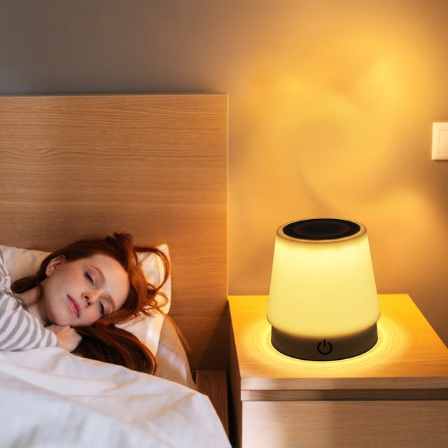 Meitianfacai small table lamp on a wooden bedside table, emitting a warm yellow glow. This USB rechargeable lamp features a compact cylindrical design with a touch-sensitive power button on its base. Suitable for bedrooms, dormitories, and study areas, it functions as a night light and bedside lamp, offering eye protection and versatile lighting for various settings.