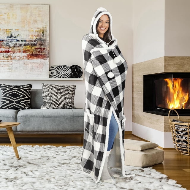 Safdie & Co. Premium Hoodzy Wearable Hooded Throw Blanket Buffalo Plaid White and Black with Sherpa worn by a person standing in a cozy living room, featuring a large black and white buffalo plaid pattern, oversized hood with pom-pom drawstrings, two front pockets, and soft sherpa lining, paired with blue jeans and white socks, near a fireplace with a burning fire, a grey sofa with black and white patterned cushions, a wooden coffee table, and a fluffy white rug. This wearable blanket is suitable for warmth and comfort and often used as a stylish accent piece in home décor.