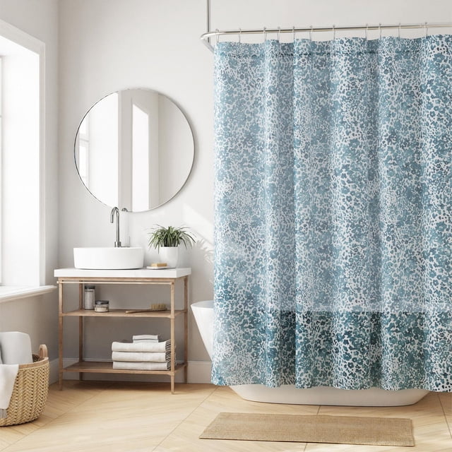 Hometrends Decorative Fabric Shower Curtain with Botanical Designs in blue and white speckled botanical pattern hanging on a curved rod in a bright bathroom with light wood flooring, round mirror, wooden vanity with white vessel sink, and woven basket with towels. Made of 100% polyester, machine washable, fits standard bathtubs.
