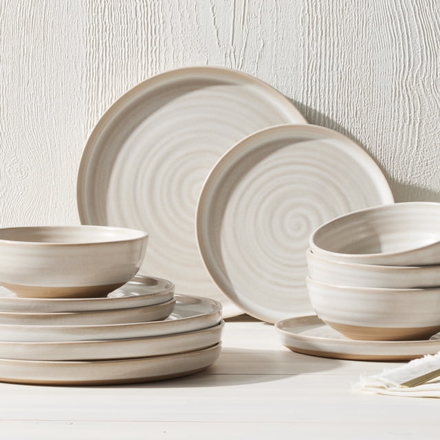 Better Homes & Gardens-Collins Cream Stoneware 12-Piece Dinnerware Set showing cream-colored stoneware plates and bowls with a reactive glaze and natural exposed clay finish, arranged on a white wood surface against a textured white wall, suitable for everyday and formal dining and often used for serving soups or salads.