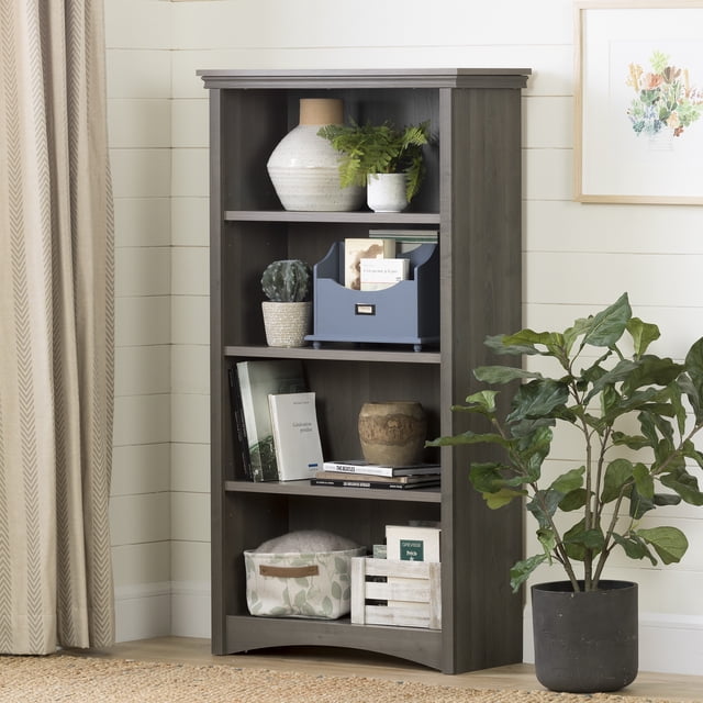 South Shore Gascony 4-Shelf Bookcase in gray maple with a contemporary style, featuring a curved kick plate and thick top molding, holding books, decorative vases, potted plants, and storage baskets, suitable for office furniture or bookshelf use in a light, neutral room with a nearby potted plant.