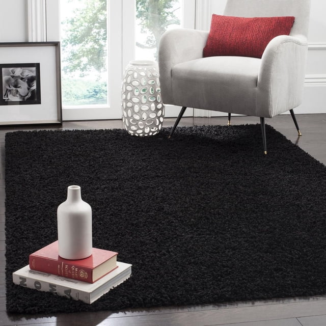 Safavieh Athens Solid Plush Shag Area Rug in black with a dense polypropylene pile, shown on dark hardwood floors. It is styled with a light grey armchair with black legs and a red pillow, a white ceramic vase on stacked books, and a white decorative ceramic stool near a window, suitable for adding plush comfort to living rooms.