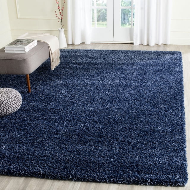 Safavieh California Solid Plush Shag Area Rug in navy blue with a dense 4.5kg/m² polypropylene pile, placed on light wood flooring in a bright room with a gray bench, books, a throw, white curtains, and glass doors, ideal for adding softness and warmth to home interiors.