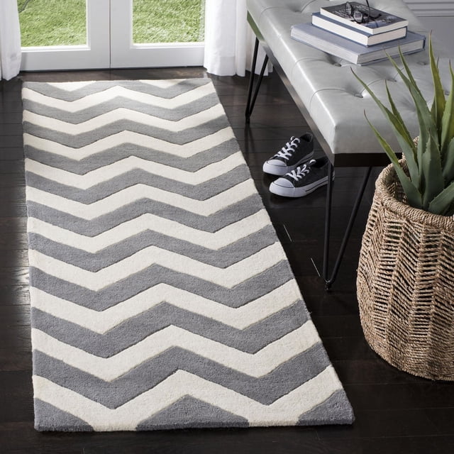 Safavieh Chatham Jake Zigzag Stripes Area Rug in a runner style with grey and white zigzag stripes and a plush medium-height wool pile, placed on dark hardwood flooring near glass doors. The rug suits contemporary entryways and hallways, shown with a grey tufted bench, books, sneakers, and a woven basket with a green plant.