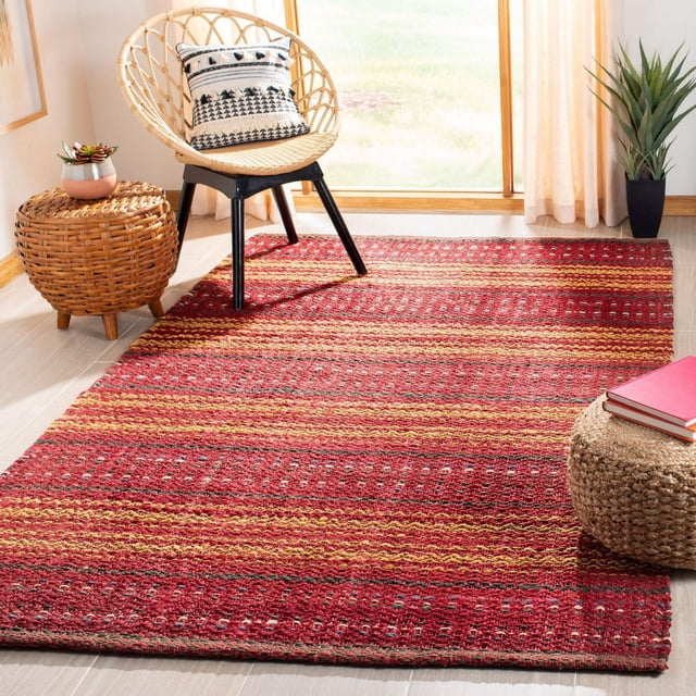 Safavieh Natural Fiber Ombre Striped Area Rug with textured red, yellow, and orange woven stripes on a light wood floor in a sunlit room featuring a rattan chair with a black and white patterned cushion, a woven side table with a small plant, and a round woven pouf with books, suitable for adding natural coastal style to living spaces.