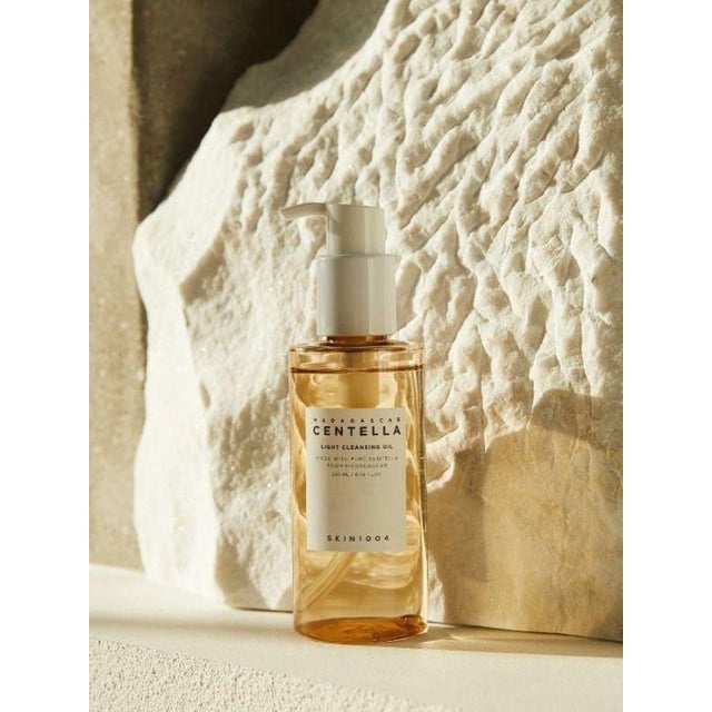 SKIN1004 Madagascar Centella Light Cleansing Oil 200ml in a transparent amber bottle with a white pump dispenser, positioned against a textured beige stone background, formulated for gentle, soothing removal of makeup, dirt, oils, and sunscreen without eye irritation, suitable for deep facial cleansing.