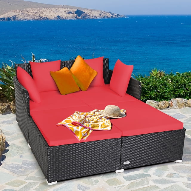 Costway Outdoor Patio Rattan Daybed with thick red cushions and four matching pillows, featuring a spacious design with armrests and a curved backrest for added comfort. Made of weather-resistant rattan with aluminum foot pads for durability, it supports up to 800 lbs. Shown on a stone patio overlooking a scenic ocean view.