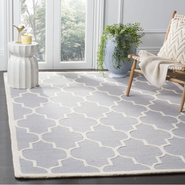 Safavieh Cambridge Kevin Geometric Area Rug in light grey with white trellis pattern, hand-tufted wool featuring plush cut pile and textured high/low loop, displayed on dark hardwood floor in a bright room with a white ceramic side table, wooden chair with cream knit throw and pillow, and a blue textured planter with green foliage, suitable for adding modern American style to home or office decor.