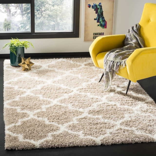 Safavieh Montreal Lennart Geometric Shag Area Rug with a beige and white plush pile in a geometric trellis pattern, placed on a dark floor beside a yellow armchair draped with a gray throw, near a window with natural light and a green plant in a dark green vase, suitable for contemporary living spaces.