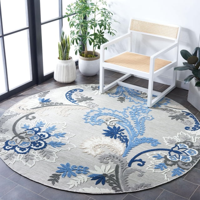Safavieh Cabana Kuzman Floral Outdoor Area Rug, round with textured synthetic yarn weave, featuring blue, gray, and cream floral patterns, placed on dark tile floor beside a white wooden chair with woven cane seat and backrest, near potted green plants by a window, ideal for outdoor patios or decks.