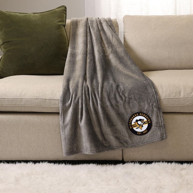 NHL, NBA, MLB Grey Sports Throw Blanket, 50" x 60", soft plush polyester draped over a beige sofa with a green cushion, featuring a Pittsburgh Penguins logo patch near the bottom corner, suitable for cozy indoor lounging or outdoor use.