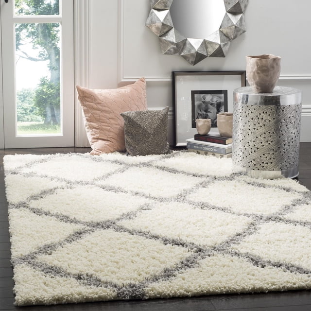 Safavieh Dallas Logan Geometric Shag Area Rug with ivory and grey Moroccan tile pattern on a soft polypropylene shag texture, shown on dark hardwood floor near a window, complemented by decorative pillows, ceramic vases, and a silver carved metal side table.