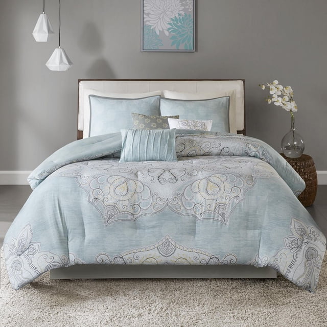 Home Essence Leslie 7pc Reversible Cotton Comforter Set on a queen bed with a white upholstered headboard. The comforter features a seafoam blue cotton sateen face with an overscale medallion print in white, grey, and yellow tones, reversible to a blue patterned cotton/polyester reverse. The set includes two standard shams with the reverse pattern, three decorative pillows in coordinating colors, and a solid grey bed skirt with a 15-inch drop. The bedroom has grey walls, two white geometric pendant lights, teal and white floral wall art, and a wicker side table with a glass vase of white orchids. This machine washable set refreshes bedroom décor with a globally inspired style.