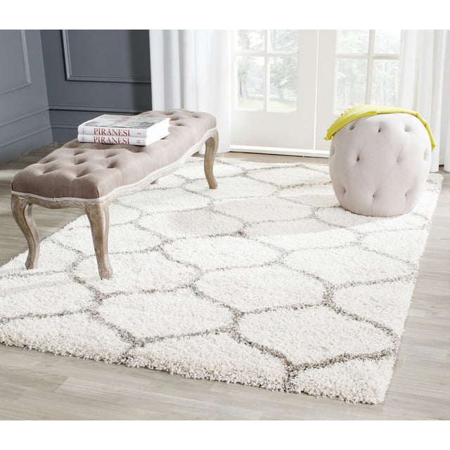 Safavieh Hudson Arline Geometric Shag Area Rug with a thick ivory shag pile and gray onion-shaped tile patterns, placed on light wood flooring in a bright room with a tufted beige bench and round beige ottoman with a yellow throw, suitable for adding texture to modern living areas.