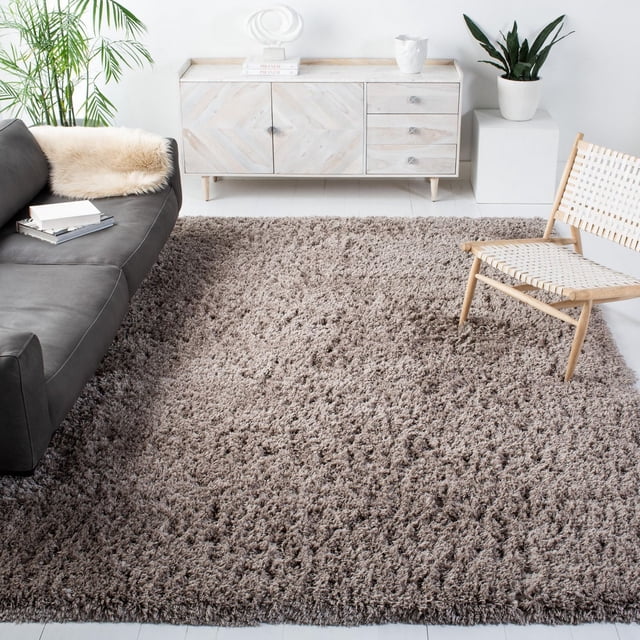 SAFAVIEH Polar Abbot Solid Extreme Plush Shag Area Rug with a 3-inch thick plush pile in warm taupe on a light wood floor, complementing a black leather sofa, woven light wood chair, and white sideboard with plants and books. The durable synthetic fiber rug adds soft texture and cozy comfort, suitable for living or relaxation spaces.