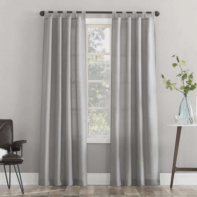 Mainstays 2-Pack Jensen Tab Top Semi-Sheer Window Curtains Panel Pair in grey, hanging on a dark brown rod with tab tops, featuring semi-sheer textured fabric that filters light and enhances privacy, displayed in a minimalist room with light grey walls, wood flooring, a black chair on the left, and a white side table with a glass vase and green leaves on the right, suitable for adding soft brightness and privacy to living spaces.