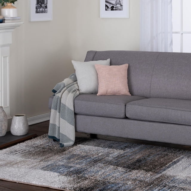 Rigton Blue and Brown Woven Area Rug with abstract navy blue, taupe, and gray tones on dark hardwood floor beneath a gray sofa with light gray and pink cushions and a striped throw. The plush rug is suitable for high-traffic areas and adds soft texture to living spaces.