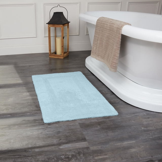 Fabstyles Basix Tapis de Bain en Coton Réversible Doux et Absorbant Bleu Pâle Placé sur un Plancher de Bois Foncé à Côté d'Une Baignoire Autoportante Blanche avec une Serviette beige Drapée au-Dessus du Bord de la Baignoire et une Lanterne en Bois avec Bougie Près d'Un Mur à Panneaux Blancs, Idéal pour une Utilisation dans les Zones de Douche Ou de Baignoire.