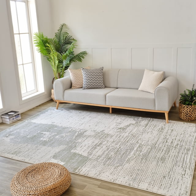 Abstract indoor area rug in green and beige tones placed in a modern living room setting with a light gray sofa, decorative pillows, plants, and wooden flooring.