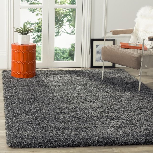 Safavieh California Solid Plush Shag Area Rug in dark grey with a dense 4.5kg/m² polypropylene pile, placed in a bright room with a glass door, an orange ceramic side table with a small plant, and a beige cushioned chair with acrylic arms and a book, suitable for adding softness and warmth to living spaces.