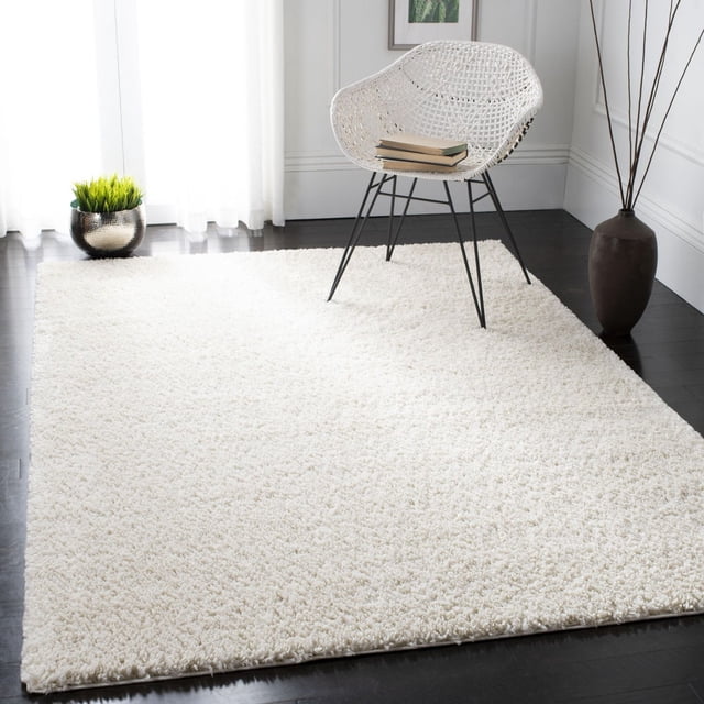 SAFAVIEH August Carlene Solid Plush Shag Area Rug in off-white on dark hardwood floors beneath a white woven chair with black metal legs holding two books. The polypropylene shag rug offers a plush texture ideal for accent rugs in living spaces. Nearby are a metallic planter with green grass and a tall brown vase with branches in a bright room with white paneled walls and sheer curtains. It is also suitable for cozy reading or relaxation areas.