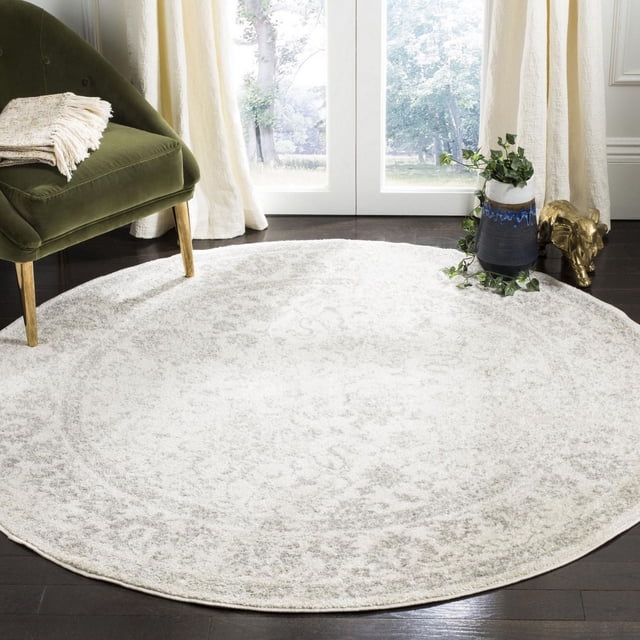 SAFAVIEH Adirondack Wyatt Traditional Area Rug, round with a subtle light grey and cream over-dye pattern, made of durable polypropylene yarns, displayed on dark hardwood flooring beside a green velvet chair and a blue and white ceramic vase with trailing ivy, in a bright room with cream curtains and glass doors, ideal for adding rustic lodge style to home interiors.