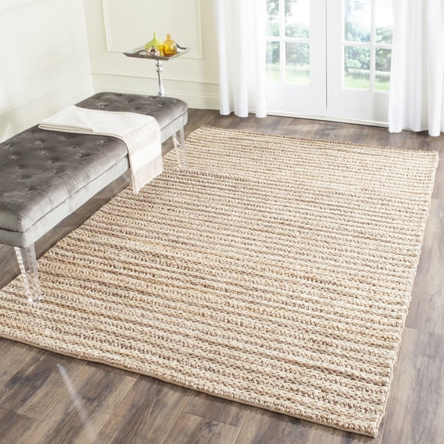 Safavieh Natural Fiber Caymen Braided Area Rug in natural beige braided sisal texture on hardwood floor, displayed in a bright room with white curtains and glass doors. A gray tufted bench with acrylic legs and a white fringed throw complements the coastal-inspired casual style.