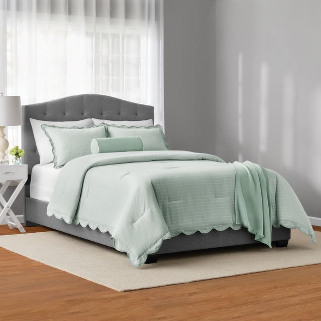 Mainstays 5-Piece Scallop Comforter Set, King, on a grey tufted bed frame in a bright bedroom. The light green comforter has pick-stitched stripes and scalloped edges, with matching king pillow shams, a decorative bolster pillow, and a soft throw at the foot. The set rests on a beige rug over wood flooring beside a white nightstand with a lamp and small green flowers. This durable, wrinkle-resistant bedding is designed for year-round comfort and easy machine washing.