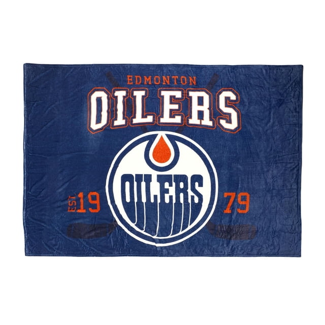 NHL Edmonton Oilers Arena Blanket, 62" x 90", a large blue fleece throw featuring the official Edmonton Oilers logo in white and orange centered. 'OILERS' appears above the logo in bold white letters outlined in orange, with 'EDMONTON' in smaller orange text above. The blanket shows 'EST 1979' in orange on each side and faint crossed hockey sticks in dark blue behind the design. This soft polyester fleece blanket is jumbo-sized for cozy warmth and team spirit indoors or at outdoor sports events.
