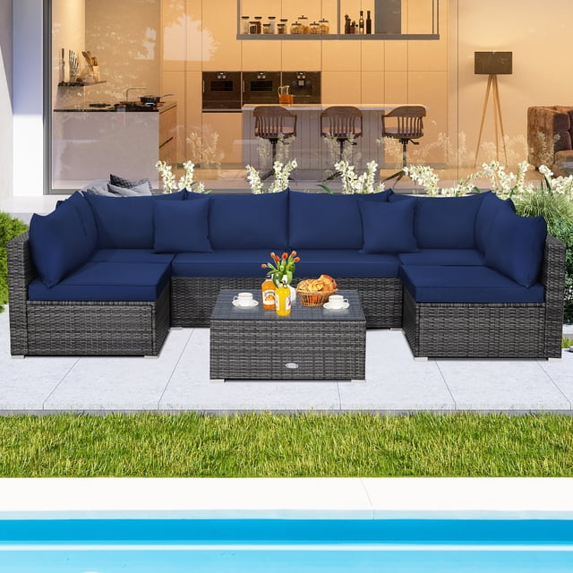 Costway 7PCS Patio Rattan Furniture Set Sectional Sofa Cushioned Garden Navy arranged in an outdoor space. Features mix grey PE rattan corner and armless sofas with navy cushions, a tempered glass coffee table, and throw pillows. Weather-resistant and ideal for patios, gardens, and poolside areas.