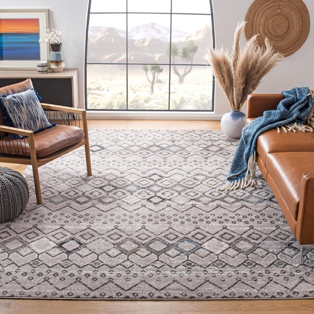SAFAVIEH Amsterdam Gladwin Geometric Area Rug featuring beige and gray tribal geometric patterns on a power-loomed synthetic yarn pile. The rug is placed on light hardwood flooring in a bright living room between a tan leather sofa with a blue throw and a wooden armchair with a blue cushion, near a large window showing a desert landscape. It is ideal for adding rustic-chic style and durable accent flooring.