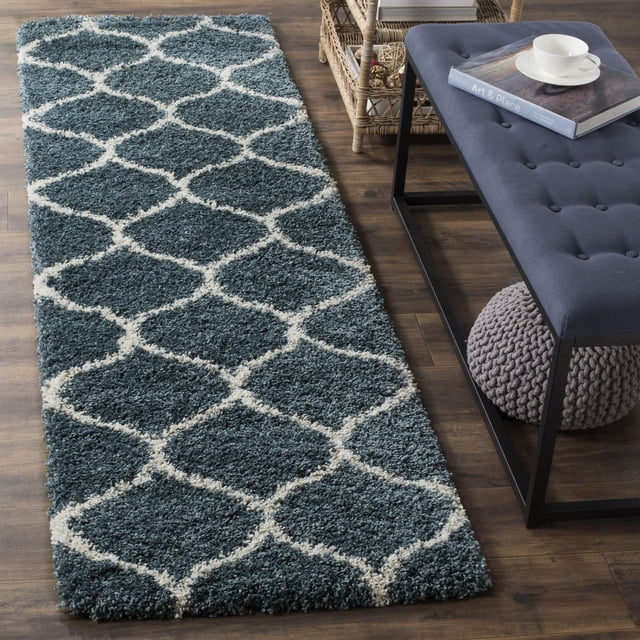 Safavieh Hudson Tapis Shag Géométrique Arline Exposé sur Plancher de Bois Franc dans une Vue Orientée vers l'Avant, Présentant une Épaisse pile Shag Pelucheuse de Couleur Sarcelle Foncée et un Motif de Carreaux Ivoire en Forme d'Oignon, Positionné à Côté d'Un Banc Capitonné Bleu Marine avec un Livre et une Tasse, Idéal pour Ajouter de la texture à un décor Moderne Ou Urbain et durable pour les Aires de Vie Très Passantes.