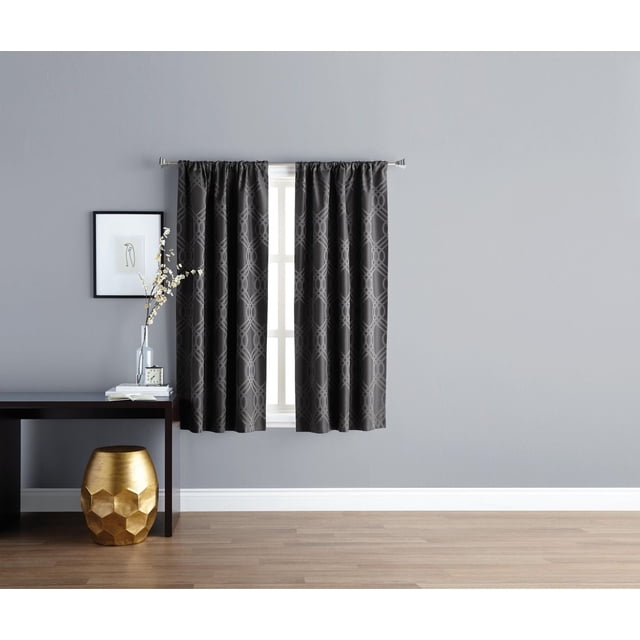 Eclipse Zana Trellis 63" Blackout Rod Pocket Window Panel in black with a wavy interlocking geometric jacquard pattern, hanging on a silver rod over a white window frame. The blackout curtain provides privacy, light blocking, and energy efficiency. It is displayed in a room with a light grey wall, dark wood console table with a glass vase of white flowers, books, and a clock, plus a gold hexagonal stool on a light wood floor.