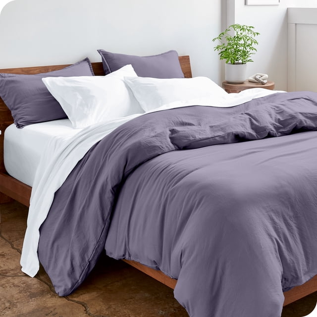 Bare Home Housse de Couette Sablée - Ultra Douce - Premium 1800 Collection - Ensemble 3 Pcs - Grand Lit, Violet Poussiéreux. l'Image Présente un Lit Habillé d'Une Housse de Couette Violet Poussiéreux et de Taies d'Oreiller Assorties, Assorti à des Draps et Oreillers Blancs. le Matériau en Microfibre Sablée a une texture Douce et Froissée, Offrant une Esthétique shabby chic. la Literie Hypoallergénique et Respirante Est Exposée dans une Chambre Lumineuse et Minimaliste avec un Cadre de Lit en Bois et une table d'Appoint Tenant une Plante