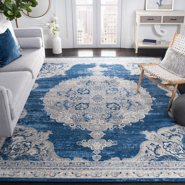 SAFAVIEH Brentwood Harland Floral Bordered Area Rug with a beige floral and geometric medallion pattern on a deep blue synthetic close cut pile, placed on dark hardwood floor between a grey sofa with a blue pillow and a wooden woven chair with a grey cushion, ideal for adding classic transitional style and warmth to living spaces.