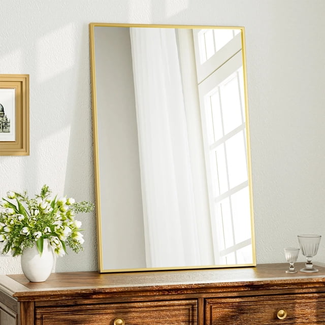 BEAUTYPEAK 24"x36" bathroom mirror with rectangular metal frame in gold, suitable for wall-mounted or leaning placement. The mirror is displayed above a wooden dresser, featuring a slim aluminum alloy frame and shatter-proof glass. It reflects a bright window and curtain, enhancing modern decor. A vase of white flowers and glassware add elegance to the setting.