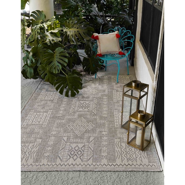ECARPET Nyra Transitional Area Rug in grey with geometric woven patterns on a textured concrete floor. Styled outdoors with a turquoise metal chair holding a cream cushion with red tassels, large green monstera leaves nearby, and two brass lanterns with white candles placed on the rug near a white wall and black window frame. Its neutral palette and texture add depth, suitable for indoor or outdoor spaces.