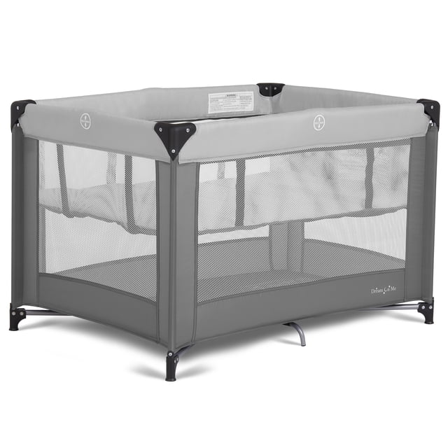 Dream On Me Zazzy Portable Play yard with Bassinet in grey, featuring a rectangular frame with mesh sides for 360-degree visibility, reinforced black corner supports, and a removable infant bassinet insert. Includes a folding mattress pad and travel bag with shoulder strap, folding compactly for easy storage and travel.