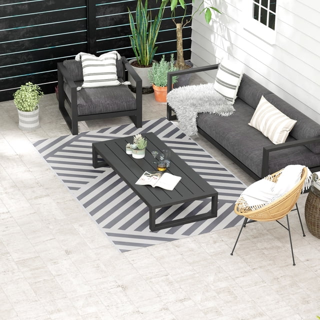 Tapis d'Extérieur Réversible pour VR, Tapis de Sol de Patio 6 Pieds x 9 Pieds, Gris et Crème, Exposé dans un Décor Moderne de Patio Extérieur. le Tapis Présente un Motif Rayé Gris et Crème, Fabriqué en Polypropylène durable avec Résistance aux UV et à l'Eau. Convient au camping, aux VR, aux patios et aux Pique-Niques, Il Est Entouré de Meubles Noirs, de Plantes en Pot et d'Une Chaise en Osier, Soulignant Sa Polyvalence.