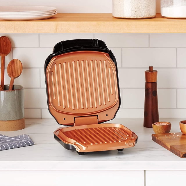 Gotham Steel Low Fat Multipurpose Sandwich Grill Nonstick Copper Coating with open copper-coloured ridged grill plates and black casing on a white kitchen counter, featuring even heating aluminum plates and a drip tray for low-fat cooking, placed near wooden kitchen utensils, a pepper grinder, and salt bowls on a cutting board.