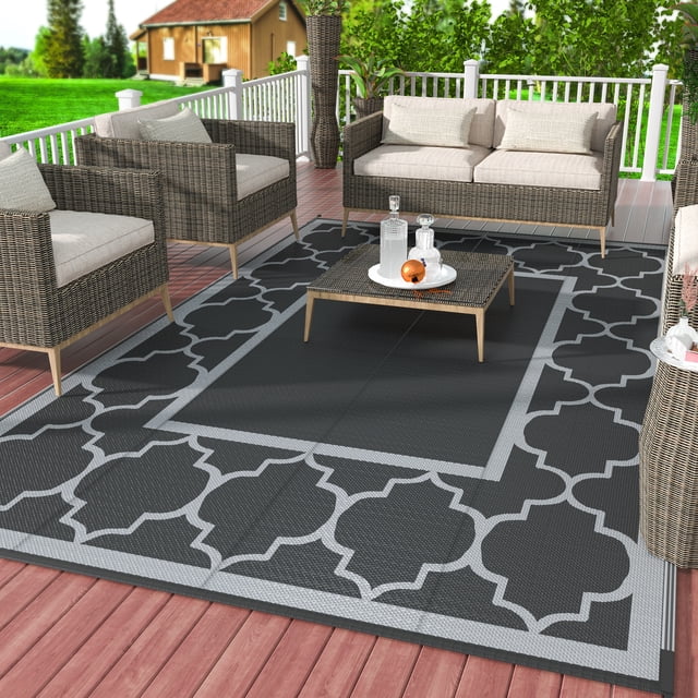 DEORAB Outdoor Rug Reversible 6x9' Patio Rug in black and grey, shown on a wooden deck with wicker furniture. The rug features a geometric pattern and is made from waterproof, UV-resistant materials for durability. Its reversible and foldable design makes it suitable for patios, camping, RVs, and other outdoor areas. Easy to clean and weather-resistant, it adds a stylish touch to any outdoor space.