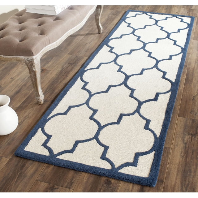 Safavieh Cambridge Kevin Geometric Area Rug with a rectangular shape, white background, and navy blue geometric pattern, hand-tufted wool texture, placed on wood flooring next to a beige tufted bench and white ceramic vase, suitable for home or office accent areas.