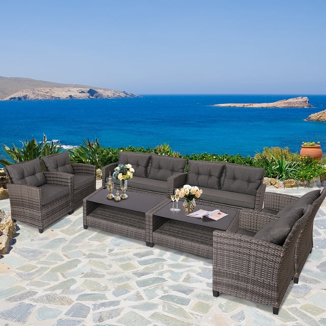 Gymax 8PCS Rattan Patio Conversation Set Cushioned Outdoor Furniture Set displayed in an outdoor setting with a scenic ocean view. The set includes two loveseats, four single sofas, and two coffee tables with tempered glass tops. The furniture is constructed with durable PE rattan and steel frames, featuring grey cushions for added comfort. The coffee tables have lower shelves for additional storage. Ideal for patios, gardens, or poolside use.